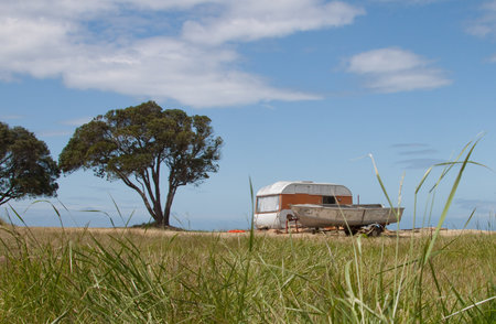 New Zealand summer freedom camping scene with caravan and aluminium fishing boat in grassy field beside a beach with trees in the backgroundの写真素材