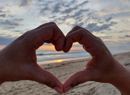 hands of a woman forming a heart and background with a beach landscape at sunsetの写真素材