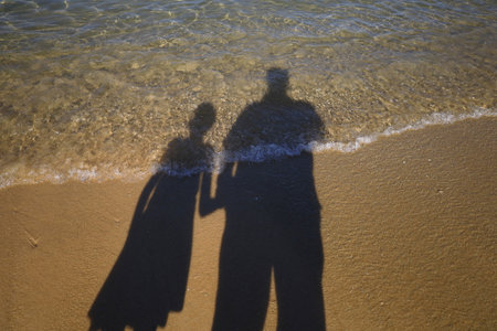 Silhouetted shadows of a father and his daughter in the reflection of the sea water and beach sandの写真素材