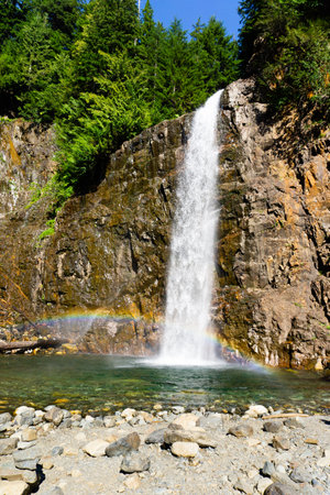 Waterfall surrounded by trees and rocks with a rainbow at the bottomの写真素材