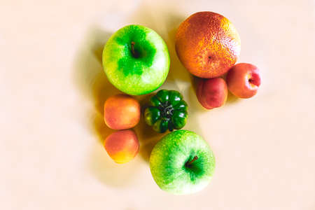 These are green apples, apricots, an orange, and a green tomato on pale beige-shaded background in a...