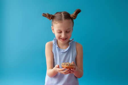 7 year old girl rejoices at the donut. She is in anticipation. She is enjoys. Hairstyle. On a blue background in a blue dress. Classic chocolate donut with sprinkles.の写真素材
