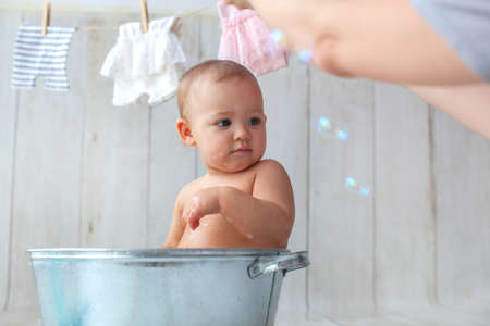 Cute baby girl takes a bath. Basin on wooden backgroun. Clothes are dried on a hanger.の写真素材