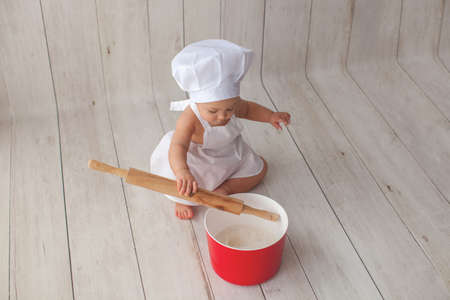 Little chef in apron and a suit of a cook, sits on a light wooden background. Chef's hat on her head. She is a bakery.の写真素材