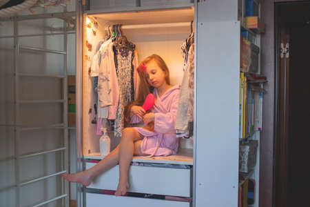 A seven-year-old girl in a pink bathrobe sits in a wardrobe and combs her long hair with a comb. Kids room interior.の写真素材