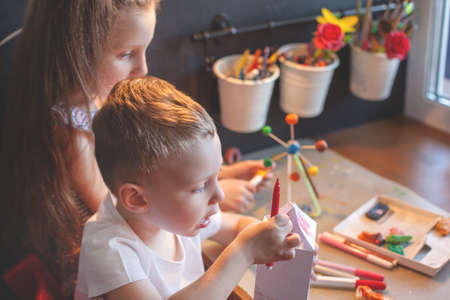 A girl of seven years and a boy of three years sit at the table and sculpt. Front view. Brother and sister. The interior of the childrens room. Chalkboard on background.の写真素材