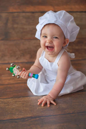 Cute one year old girl in apron is sitting in the photo zone. Birthday photoshoot and smash cake party. Portrait of little chef. The girl holds a rattle in her hands and smiles.の写真素材