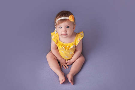 One year old girl in a yellow bodysuit sits on a blue background. Smash cake concept. Flower on her head.の写真素材