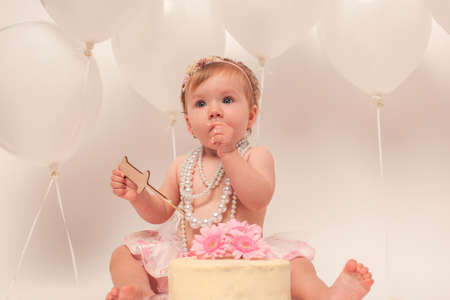One year old girl celebrating birthday. Smash cake concept. Decoration with white balloon. Flower. Beads.の写真素材