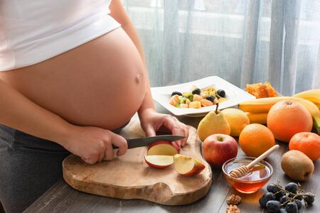 Young and beautiful pregnant woman preparing healthy fruit salad at home. Cropped image of pregnant woman cooking a fruit salad with kiwi, banana, apple, pear, tangerine, honey and walnut.の写真素材