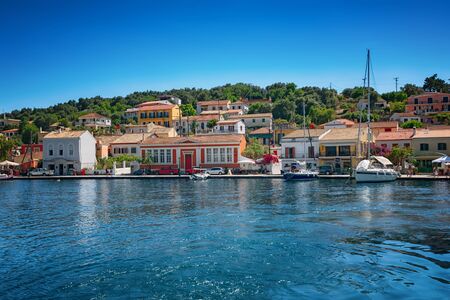 Paxos Island, Greece. View of beautiful Loggos Harbor - sea bay with calm turquoise water, ships and yachts colorful old houses and blue sky with white clouds. Summer cityscape.の写真素材