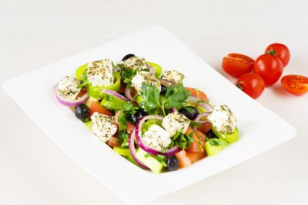 Greek salad with fresh tomato, cucumber, red onion, basil, feta cheese, black olives, Italian herbs and olive oil in white dish on white wooden table.の写真素材