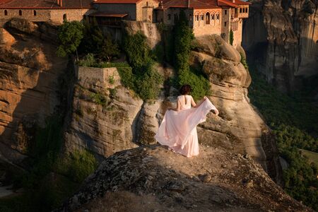 Beautiful young woman in pink dress is standing on the rocks in front of the Varlaam Monastery. Sunset light. Pindos Mountains, Thessaly, Greece, Europeの写真素材