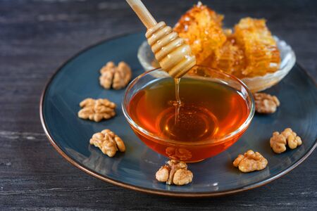 Image of honey dripping from honey dipper in glass bowl. Thick honey dipping from the wooden honey spoon, closeup. Top view of honey dipper, honey comb, walnuts and glass bowl with honey in grey dish.の写真素材