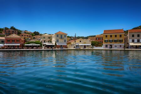 Paxos Island, Greece. View of beautiful Loggos Harbor sea bay with calm turquoise water, ships and yachts colorful old houses and blue sky with white clouds. Summer cityscape.の写真素材