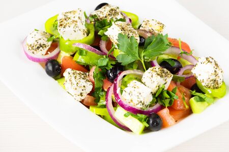 Greek salad with fresh tomato, cucumber, red onion, basil, feta cheese, black olives, Italian herbs and olive oil in white dish on white wooden table.の写真素材