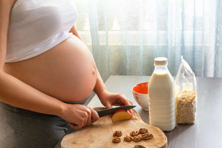 Young and beautiful pregnant woman preparing healthy breakfast at home. Cropped image of pregnant woman cooking a breakfast with oat flakes, apple, milk and walnut.の写真素材