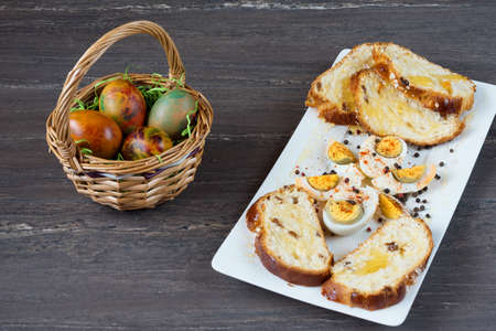 Easter wicker basket with colored eggs and sliced Easter bread in white plate on grey wooden board.の写真素材