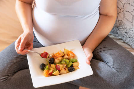 Pregnant woman eating healthy homemade organic fruit salad sitting on her bed. Close up on tummy....
