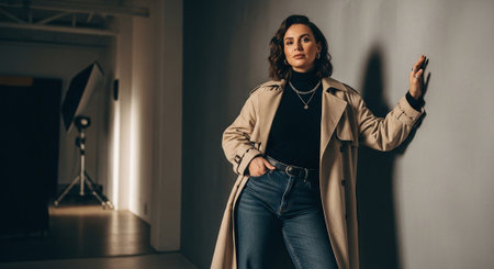 A stylish woman in a beige trench coat and black turtleneck, posing against a gray wall in a modern studio settingの素材