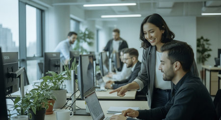 A modern office environment with professionals collaborating, a woman pointing at a laptop screen while a man looks on, greenery in the background, and colleagues working at desksの素材