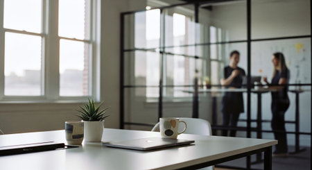 A modern office setting with a focus on a desk featuring a laptop, a plant, and coffee mugs, while two people are seen in the background having a discussionの素材