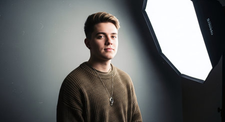 A young man with short hair wearing a knitted sweater, standing in front of a large softbox light, with a neutral backgroundの素材