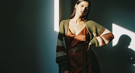 A woman posing in a stylish outfit with a knitted cardigan and a satin dress, illuminated by soft light, creating a dramatic shadowの素材