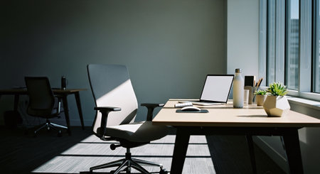 A modern office space featuring a desk with a laptop, a chair, and a plant, illuminated by natural light from large windowsの素材