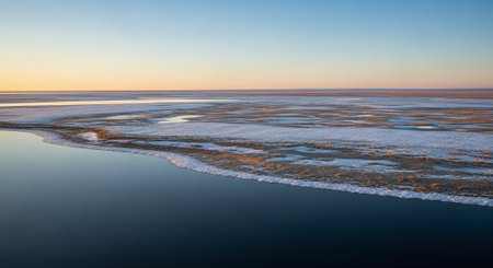 A serene landscape featuring a frozen lake with patches of ice and snow, reflecting the soft colors of a sunset skyの素材