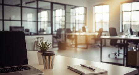 A modern office workspace featuring a laptop, a coffee cup, a small plant, and a notepad on a desk, with large windows allowing natural light to illuminate the spaceの素材