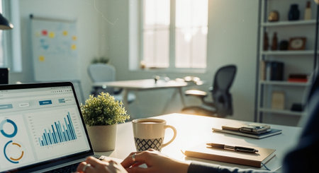 A person working on a laptop in a modern office, displaying graphs and charts on the screen, with a coffee cup and a plant on the deskの素材