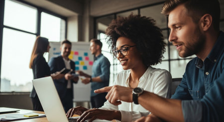 A diverse group of professionals collaborating in a modern office, with a focus on a woman working on a laptop and a man pointing at the screen, while others discuss in the backgroundの素材
