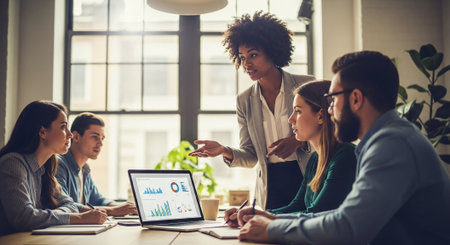 A diverse group of professionals in a modern office setting engaged in a business meeting, with a woman presenting data on a laptop, charts and graphs visibleの素材