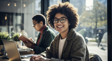 Portrait of young afro american woman in eyeglasses smiling and looking at camera while working with laptop in officeの素材