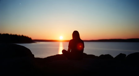 A person sits on rocks by the water during a vibrant sunset, creating a serene and contemplative scene.の素材