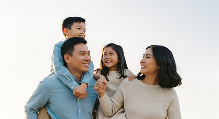 A joyful family of four, including a man carrying a young boy, posing outdoors with smiles on a bright day.の素材