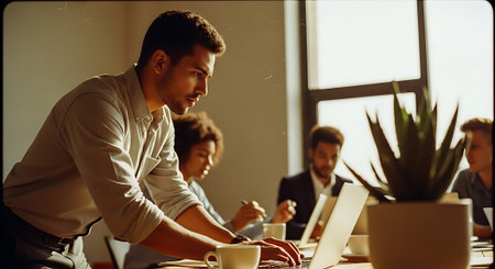 A group of professionals collaborating in a bright office space, using laptops and notebooks, with a large plant and natural light.の素材