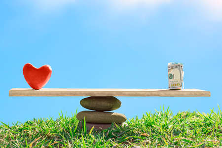 Red heart and dollar bills on the scales of stones and board on a blue sky background. The balance between love and money. life or career. Family or career choice. health care concept.の写真素材