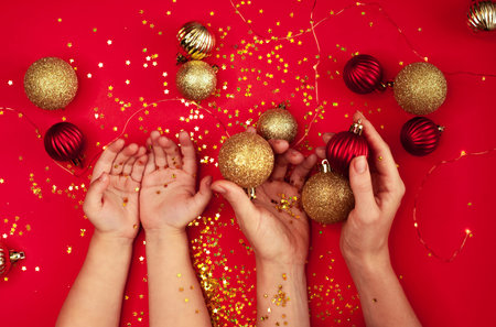 Baby and mom hands, holding Christmas decorations: golden and red balls, bright star sparkles on red...