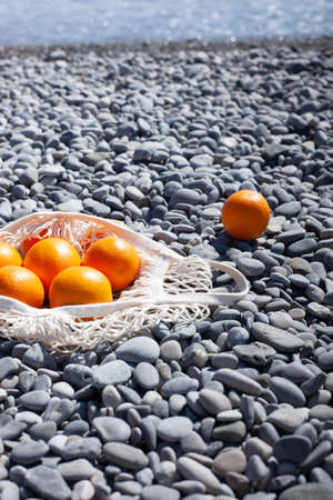 White cotton eco string bag with oranges on a rocky beach. Sea view with a blue sky. Zero and eco waste. No plastic. Bright background with copyspace. Closeup.の写真素材