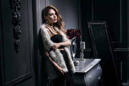 Sexy young woman in black dress and white fur jacket posing in vintage dark interiorの写真素材
