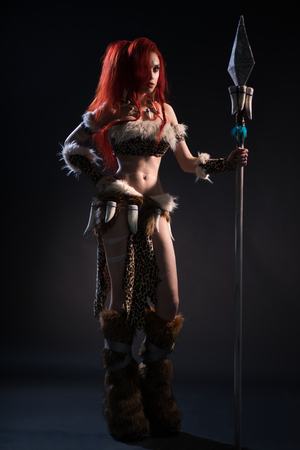 Beautiful red haired woman in stone age clothing with spear posing at camera on dark backgroundの写真素材