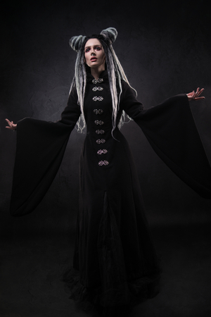 Full length view of woman with dreads wearing black gothic coat and posing on dark backgroundの写真素材