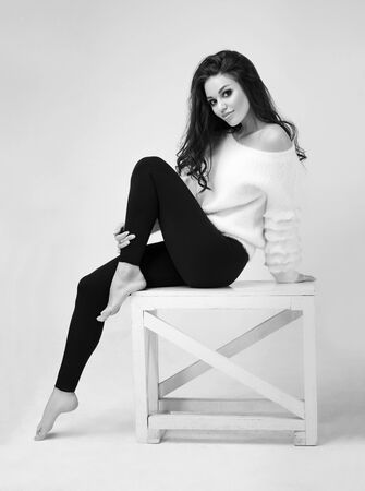 Brunette woman in casual clothes posing while sitting on wooden table against light studio backgroundの写真素材