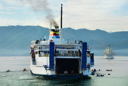 Asian overcrowded recycled ferry approaching port at dawn after an overnight journey with passengers anxious to debark and surrounded by beggars in small canoes. Mountains in the background. Cagayan De Oro City, Mindanao, Philippines.の写真素材