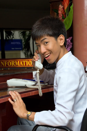 Asian smiling and welcoming hotel receptionist boy holding telephone hook or calling sitting at the desk of a hotel lobby with an information sign.の写真素材