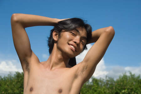 Portrait of a happy and smiling shirtless Asian teenager stretching against an azure blue sky.の写真素材