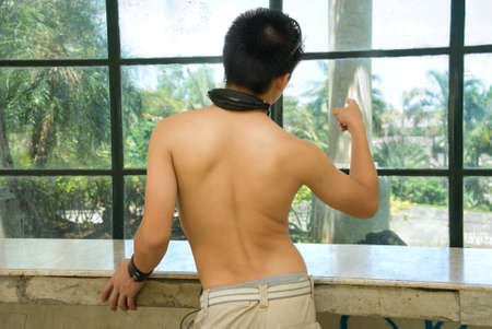 Shirtless male teenager in jeans and  with headset on shoulders pointing to a tropical cityscape through a dirty urban window, back view.の写真素材