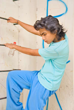 Greek teenager boy with wavy black hair in jogging outfit and with water bottle in pensive mood hanging on a ladder in an urban environment with graffiti.の写真素材